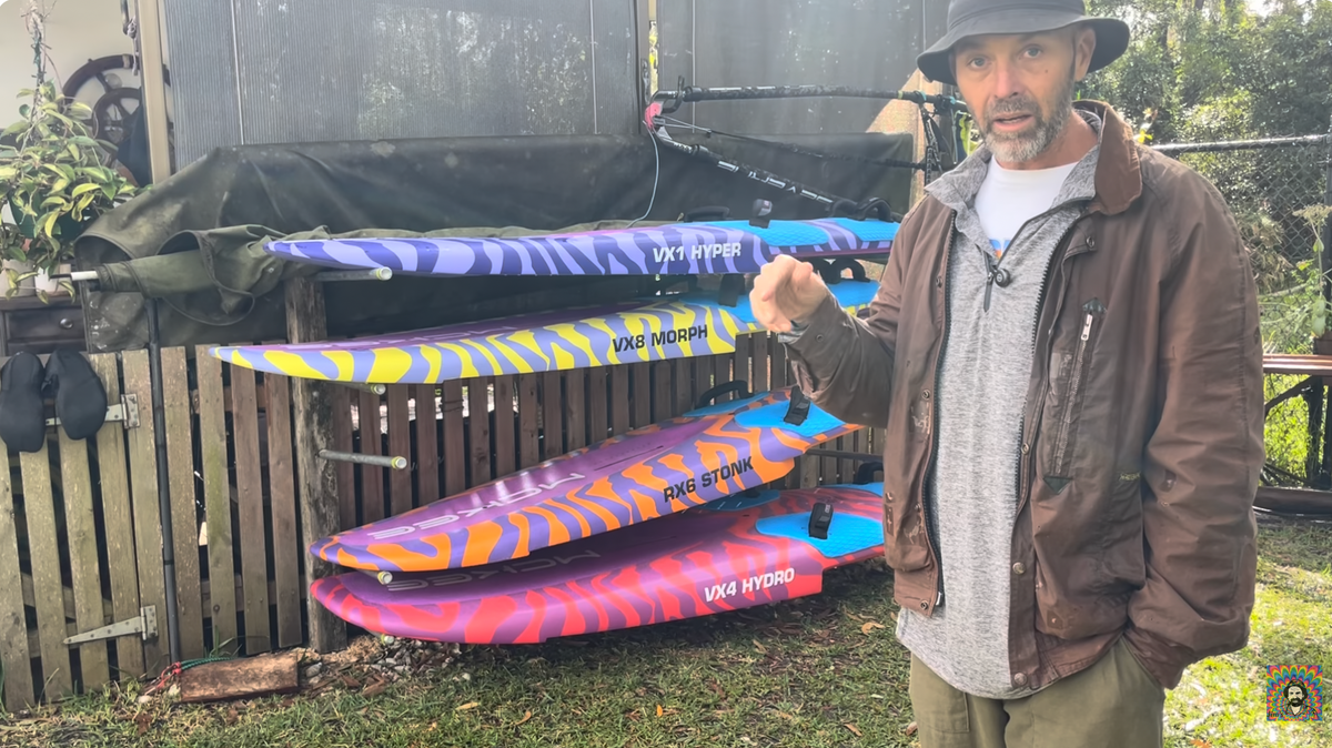 A man wearing a hat, brown jacket, and grey shirt stands next to a stack of colorful Vibe radically new Hydroplaning windsurf hulls in an outdoor backyard setting right after trying them out on the water